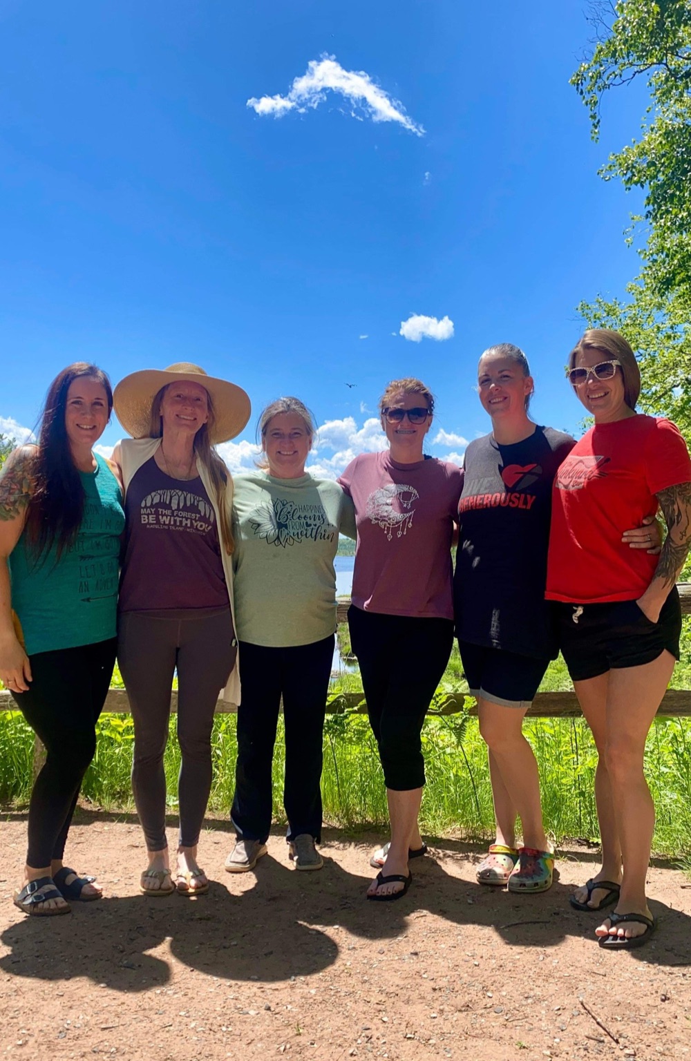 Six women standing arm in arm in the sun with Lake Superior and blue sky behind them