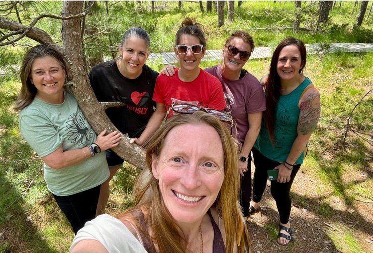 Six women smiling for a group selfie among the pines on Madeline Island