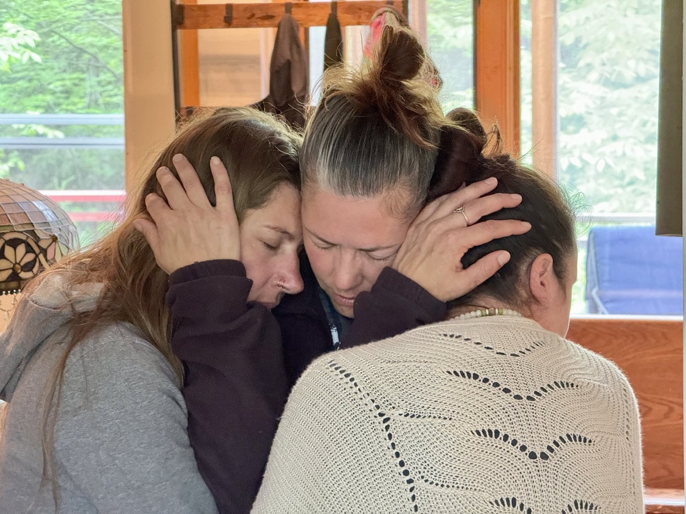 Three women holding each other close in a quiet embrace beside a sunlit window